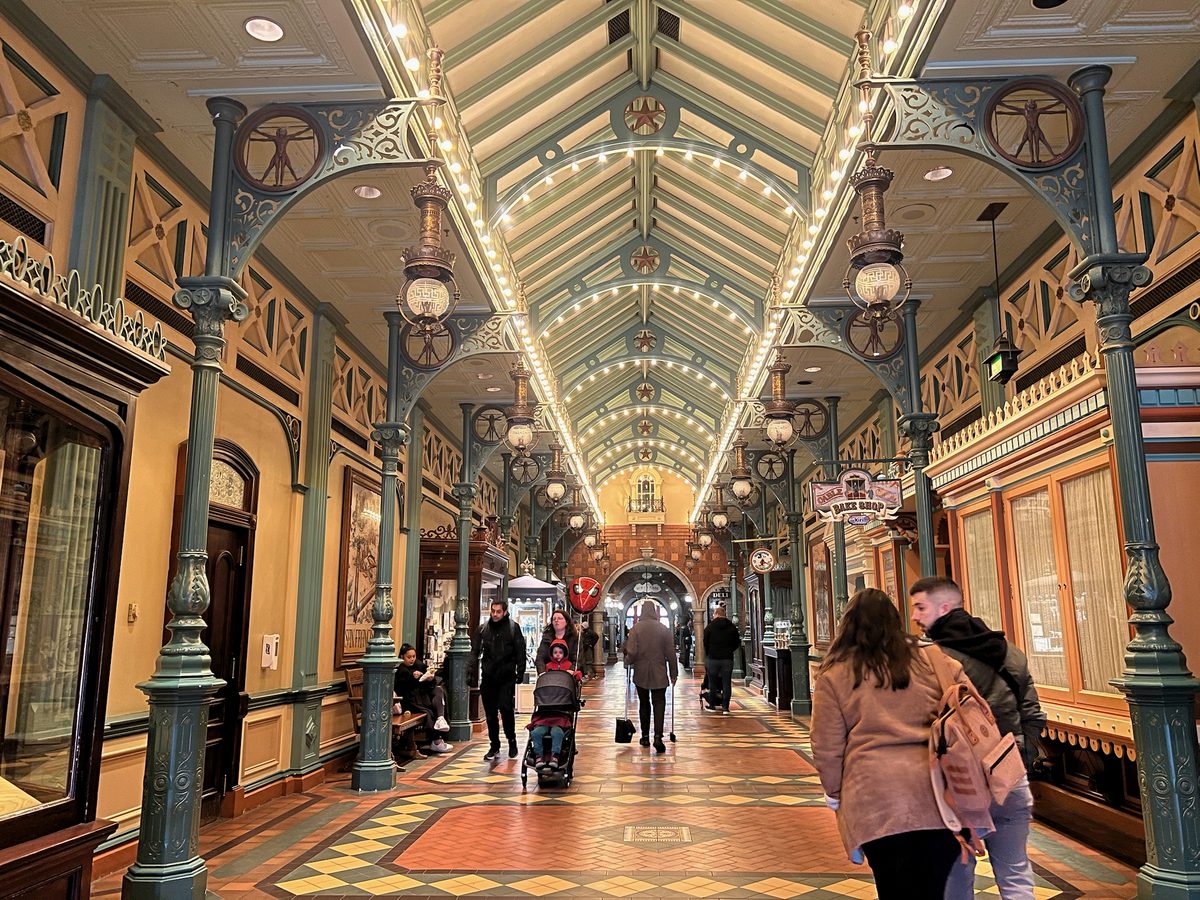 Families with a pushchair walking the tiled floor of a covered arcade beside Main Street