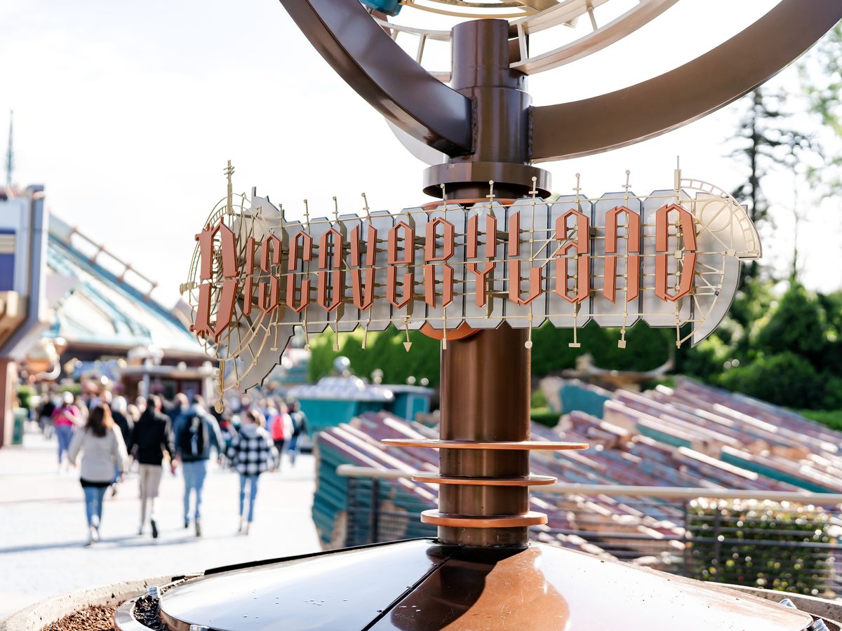 The bronze Discoveryland entrance sign on a rotating orrery-style post, with the land’s rooftops and a rollercoaster track behind