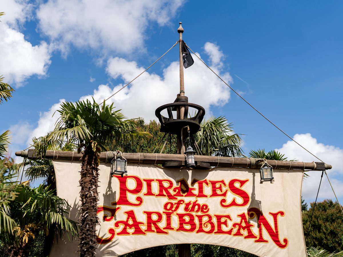 The Pirates of the Caribbean ride entrance sign, a ship’s mast and sail with the ride name in red lettering, framed by palm trees