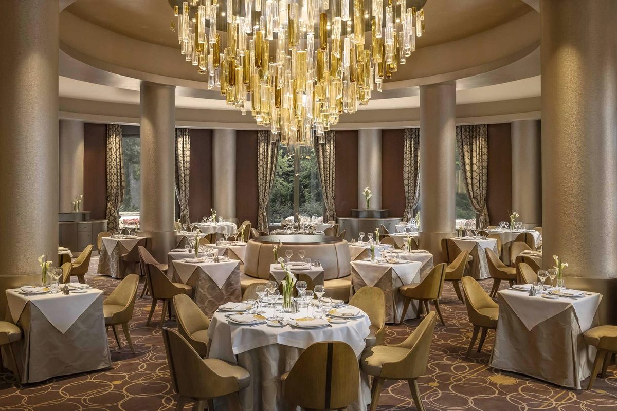 The dining room of Manhattan Restaurant, round columns and white-clothed tables beneath a large amber glass chandelier