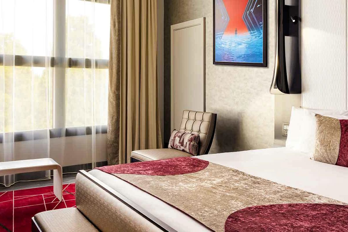 A guest room with two double beds, red and gold crushed-velvet runners, a Barcelona-style chair and a single framed Marvel print above the bed