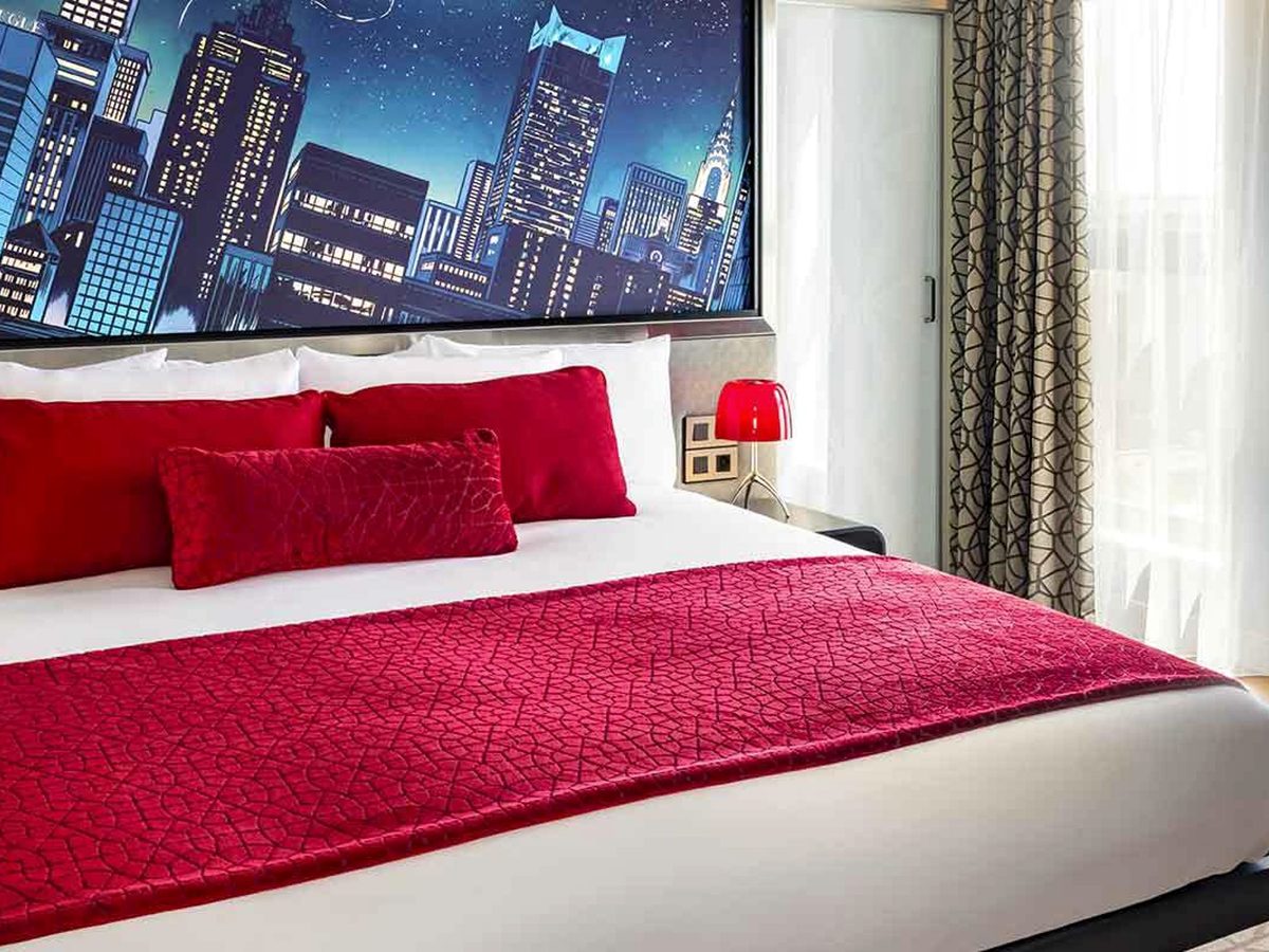 The Spider-Man Suite bedroom, a red-dressed bed beneath a wall-wide mural of a night-time Manhattan skyline