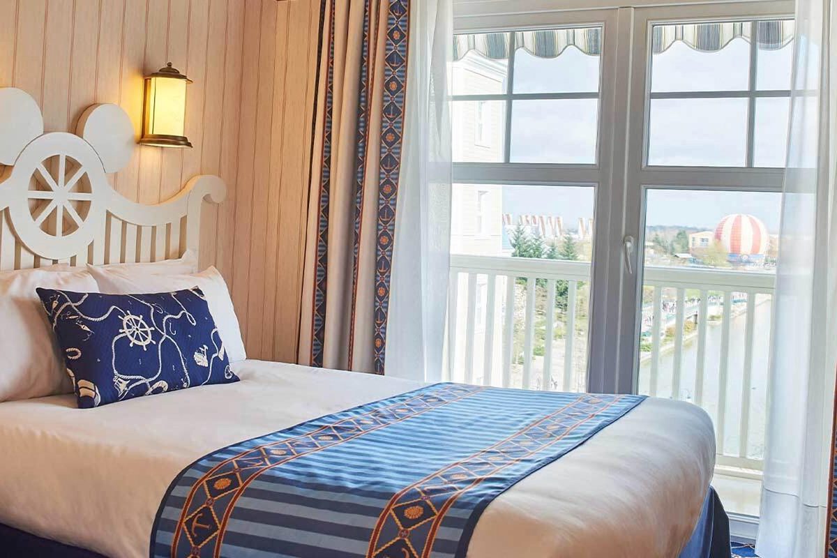 A lake-view room at Disney Newport Bay Club, a white Mickey ship-wheel headboard beside French windows onto a Juliet balcony, with Lake Disney and the Disney Village balloon beyond