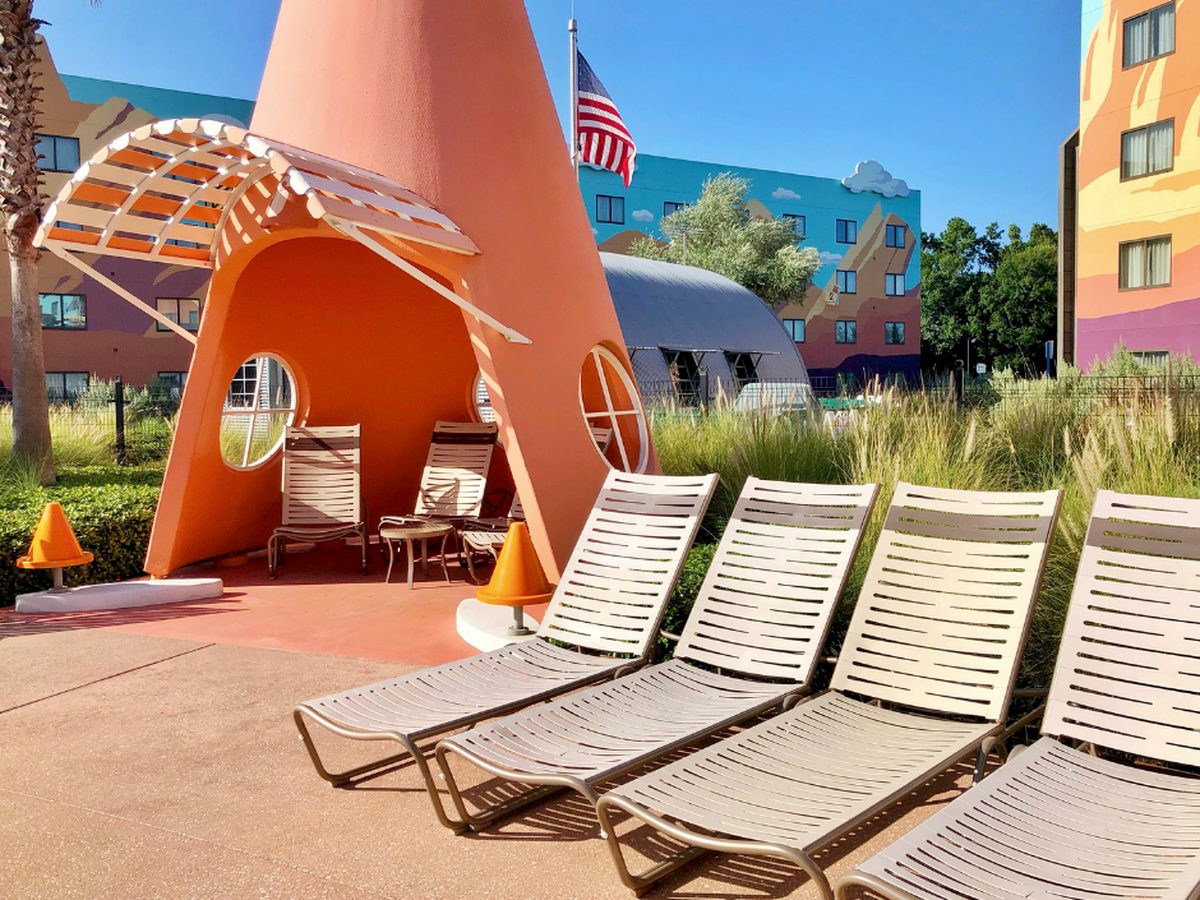 A traffic-cone-shaped cabana sheltering chairs beside sun loungers at the Cozy Cone Pool at Art of Animation, with the painted Cars buildings behind