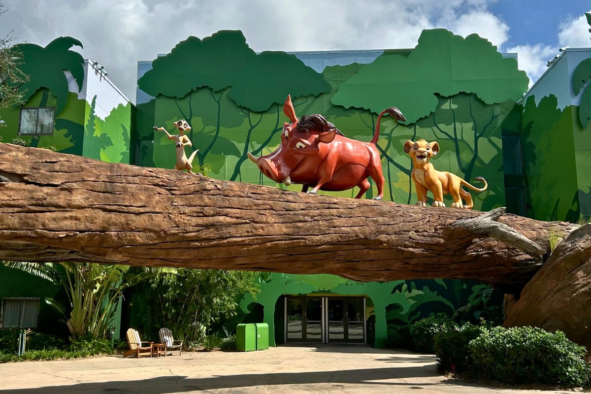 Statues of Timon, Pumbaa and young Simba crossing a fallen log against a green jungle-painted building in the Lion King courtyard at Art of Animation