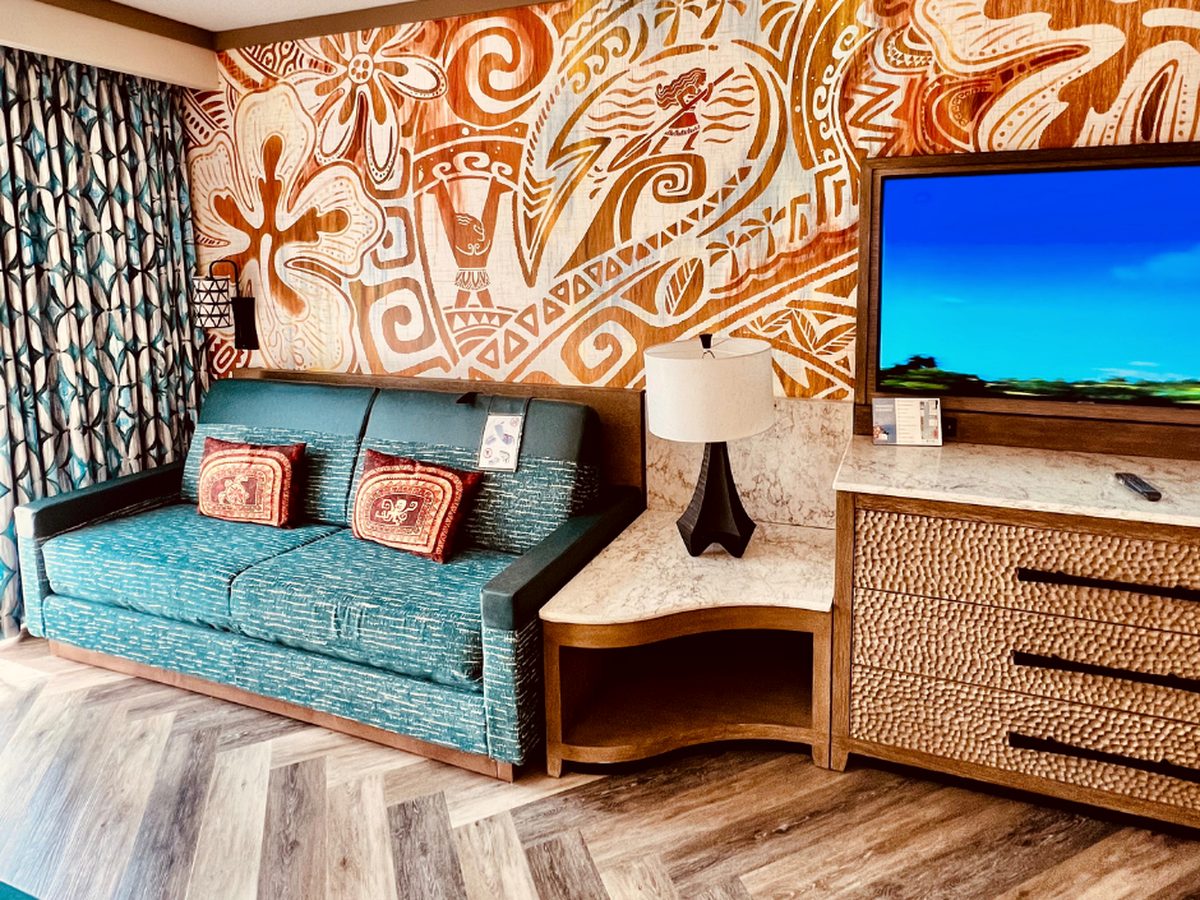 The sofa end of a guest room at Disney's Polynesian Village Resort, a teal sofa with patterned cushions beneath an orange-and-cream Moana wall mural, beside a pebble-fronted dresser and television