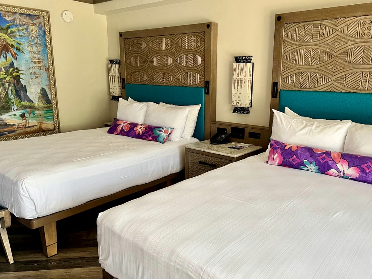 A standard room at Disney's Polynesian Village Resort with two queen beds, carved wooden headboards over teal panels, purple floral bolster cushions and framed Moana artwork