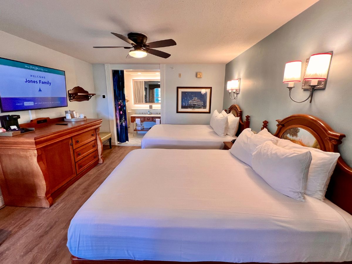 A standard room at Disney's Port Orleans Riverside, two queen beds with wooden headboards, a dresser and a television showing the resort welcome screen