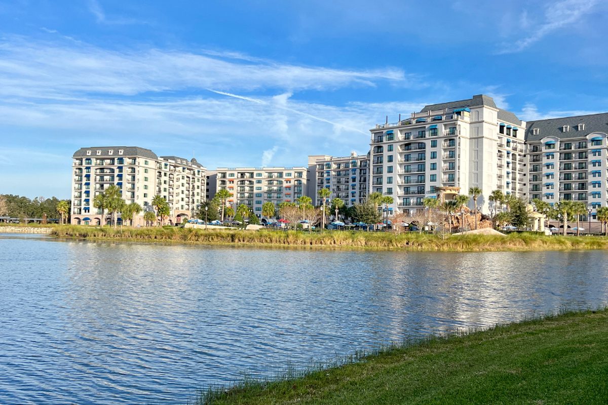 The white wings of Disney's Riviera Resort with grey roofs and balconies, seen across a lake under a blue sky