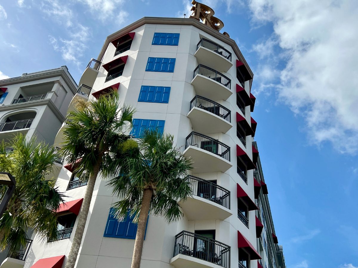 A turreted corner of Disney's Riviera Resort with blue shutters, red awnings and a gold monogram on the roof, behind two palm trees