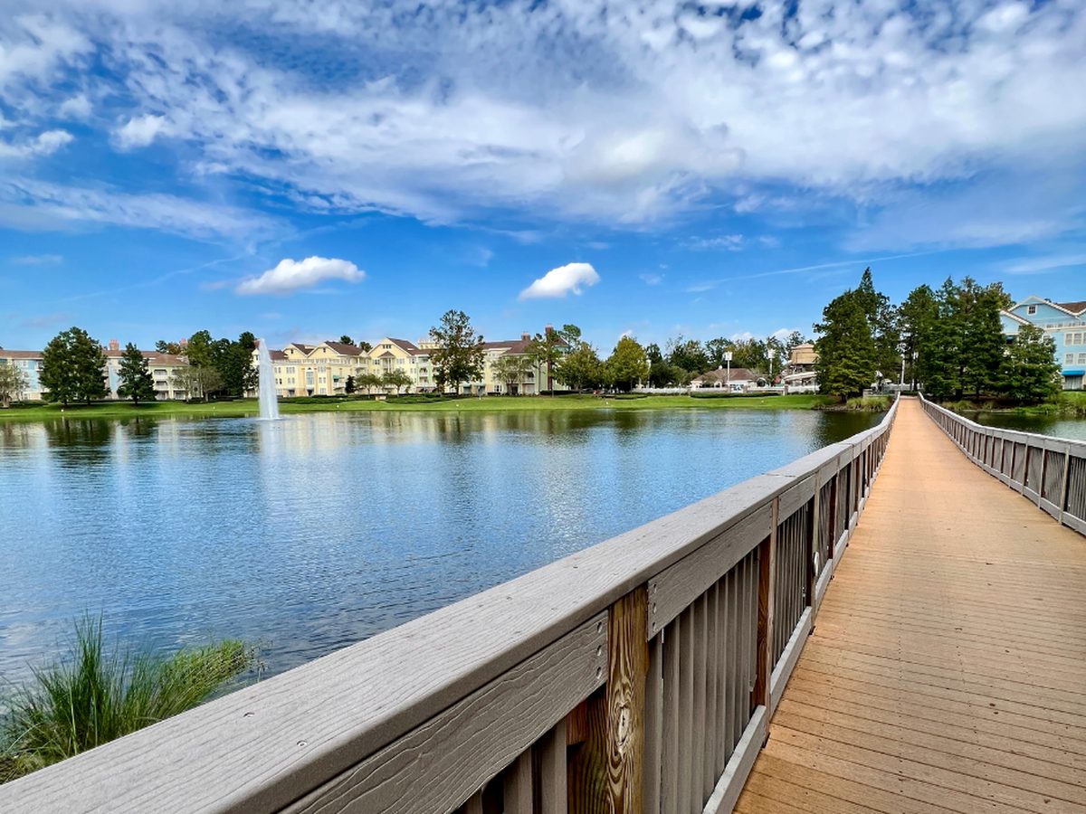 A long wooden footbridge crossing the lake at Disney's Saratoga Springs Resort, with pastel villa buildings and a fountain on the far shore under a blue sky