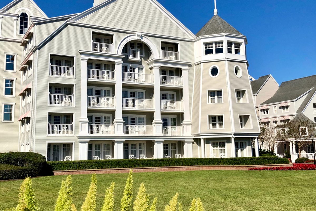 The grey clapboard facade of Disney's Yacht Club Resort, five storeys of balconied rooms and an octagonal turret above a lawn under a clear blue sky