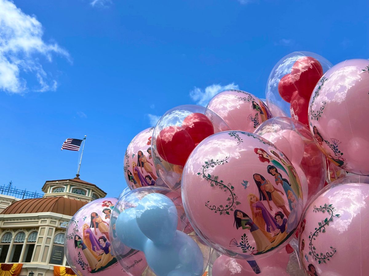 A bunch of clear balloons printed with Disney princesses, red balloons inside them and a pale blue Mickey-shaped balloon, for sale on Main Street, U.S.A. at Magic Kingdom with a US flag on a rooftop behind