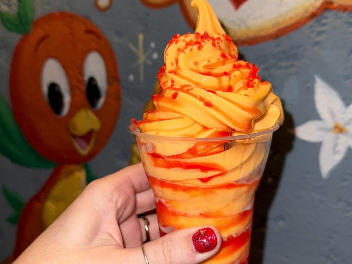 A frozen orange swirl treat in a cup held up in front of the Orange Bird mural at Sunshine Tree Terrace, Magic Kingdom