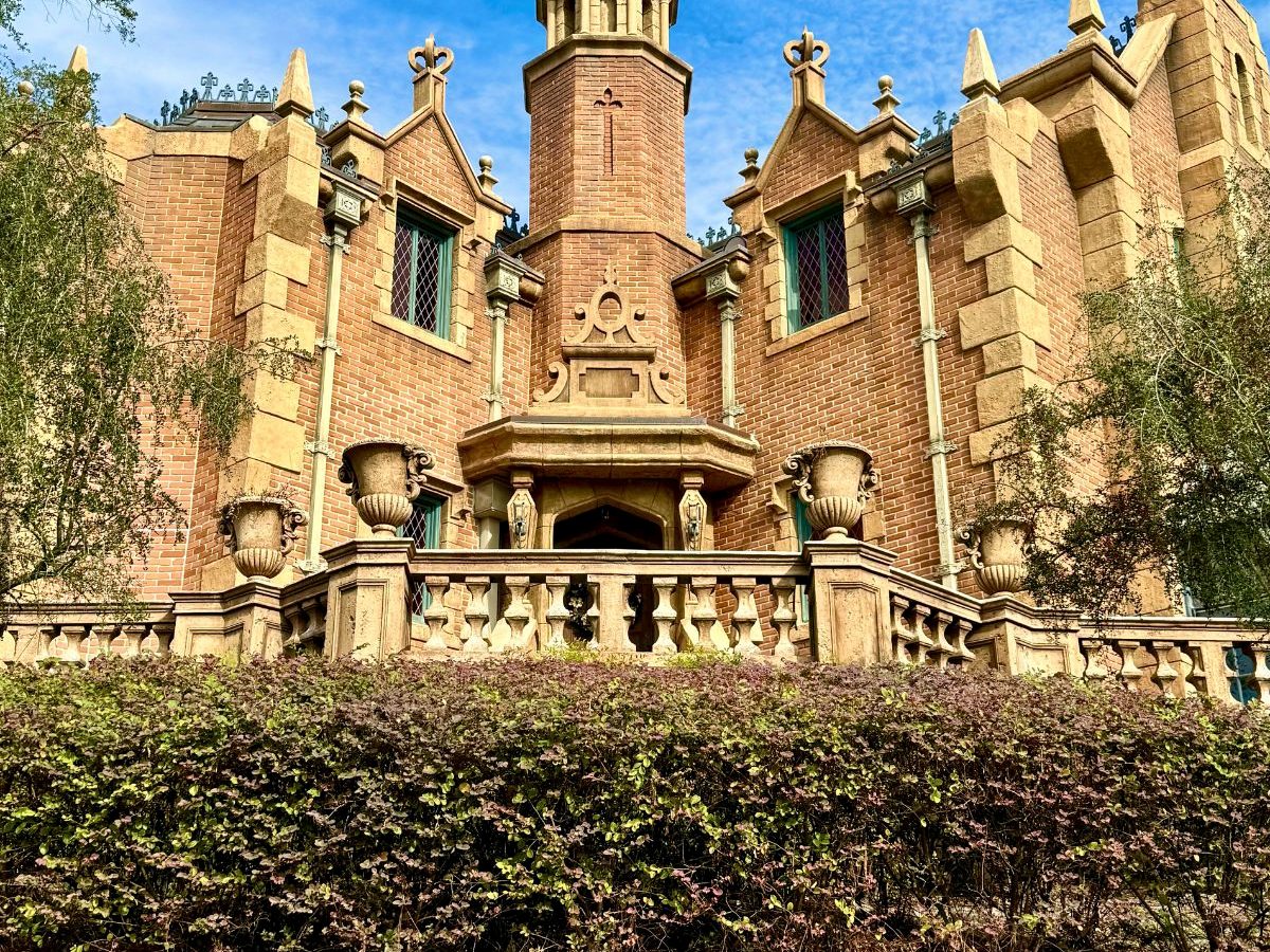 The red-brick gothic Haunted Mansion behind its lawn and hedges in Liberty Square at Magic Kingdom, under a blue sky