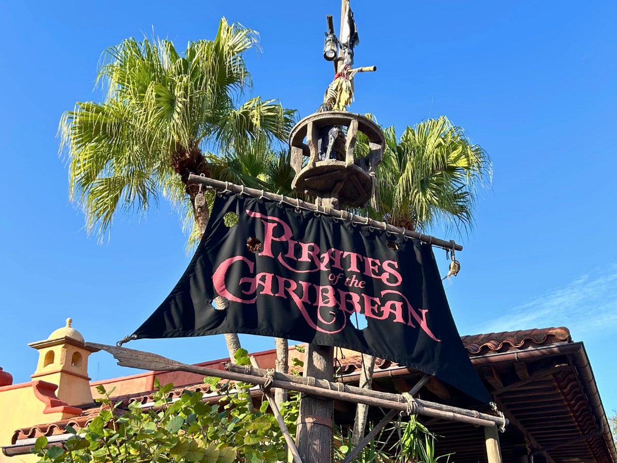 The black Pirates of the Caribbean sail sign above the Adventureland entrance at Magic Kingdom, with a crow's nest and palm tree behind against a blue sky