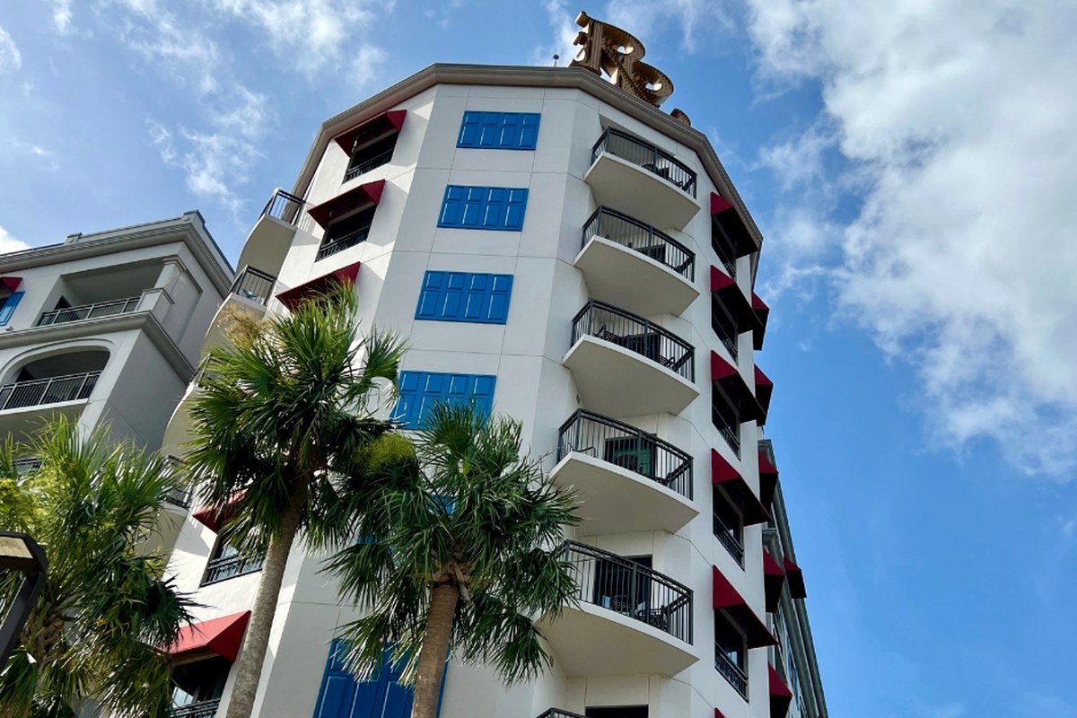 The white turret of Disney's Riviera Resort with blue shutters, red awnings and a gold rooftop monogram, palm trees in front and a blue sky behind