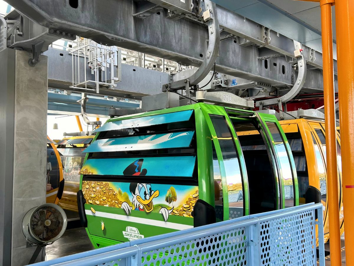 A green Disney Skyliner gondola with Scrooge McDuck artwork waiting in a station loading bay, with yellow and orange cabins behind