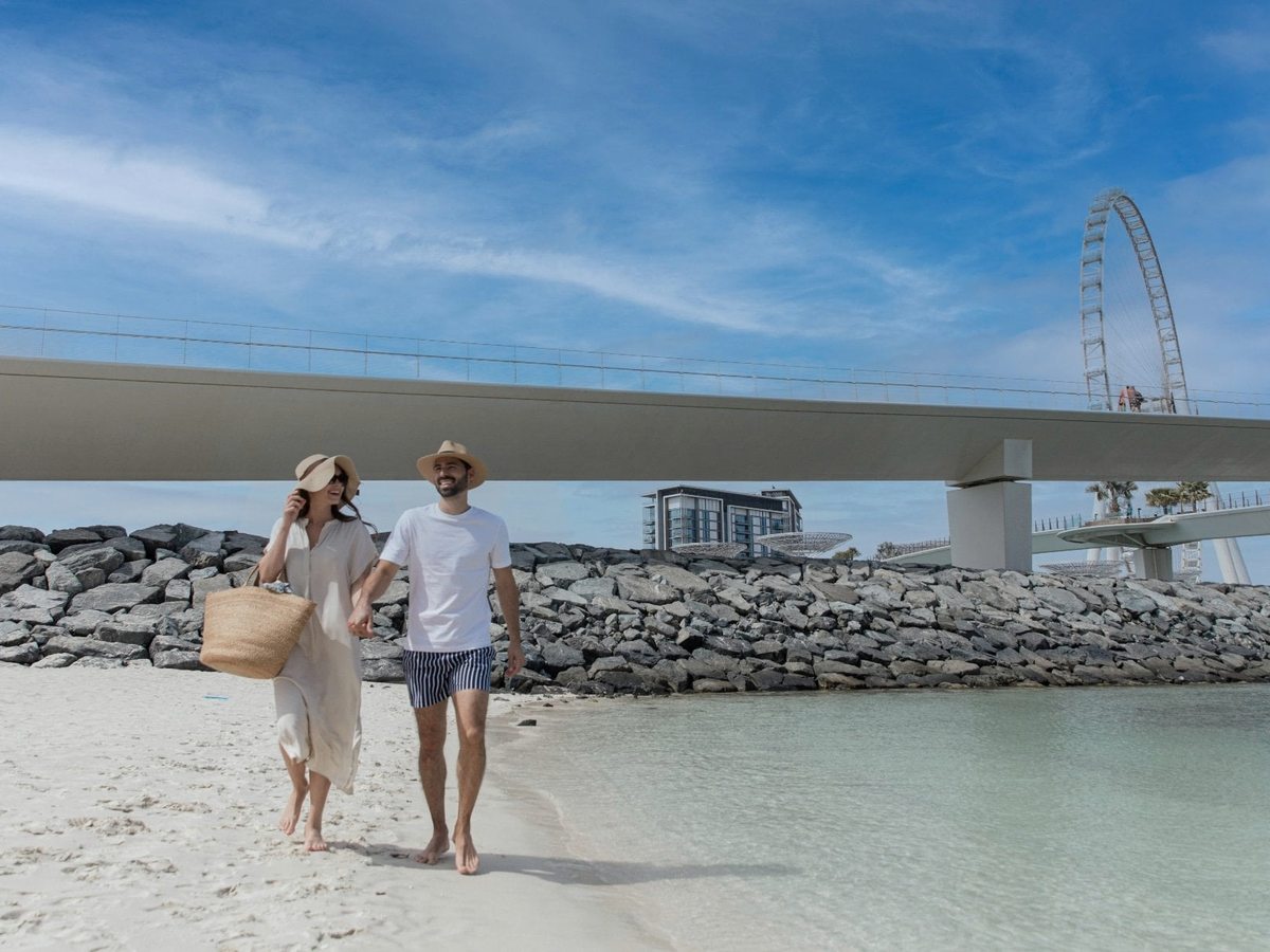 A couple walking at the water's edge on soft sand, with a breakwater, the Ain Dubai wheel and a footbridge on the far shore.