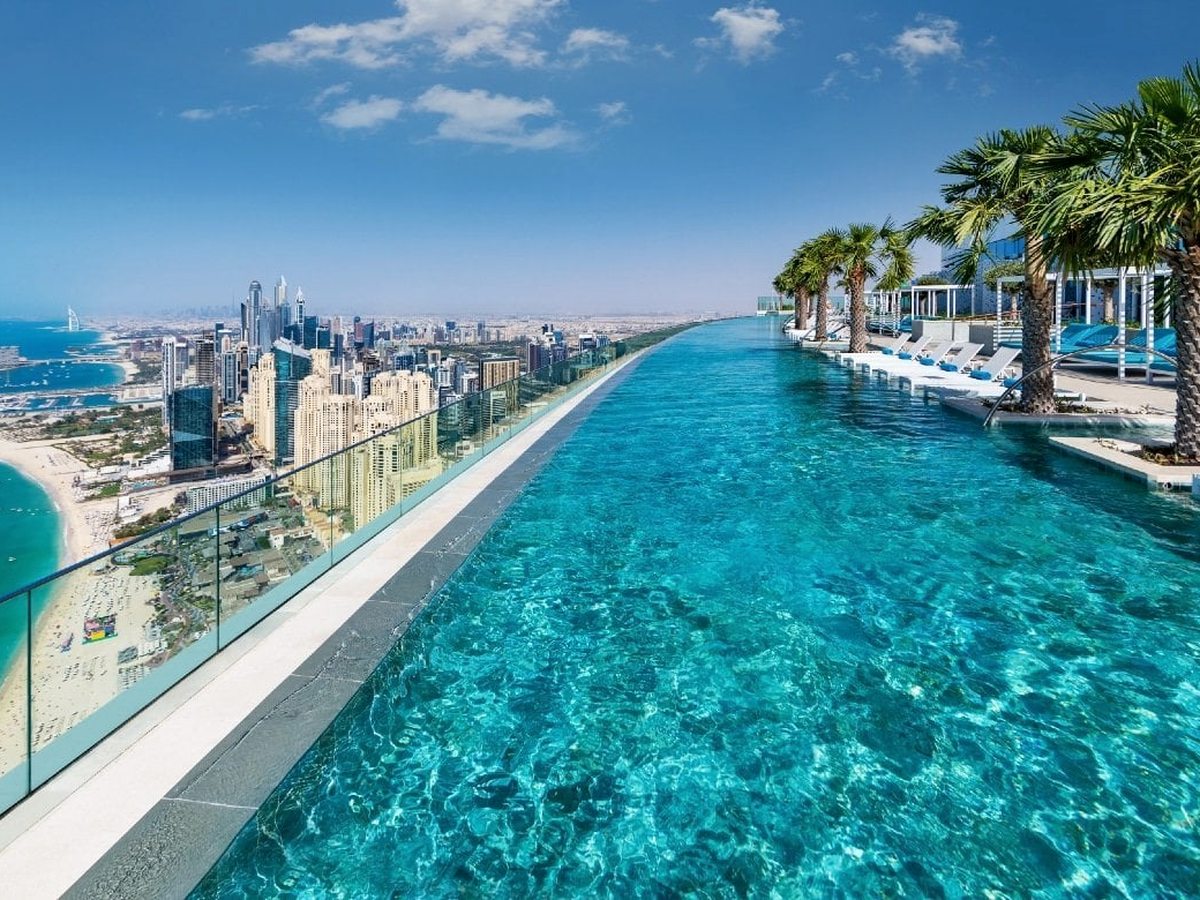A long rooftop infinity pool stretching along the tower edge, its surface meeting the horizon, with the Dubai coastline and Ain Dubai far below.