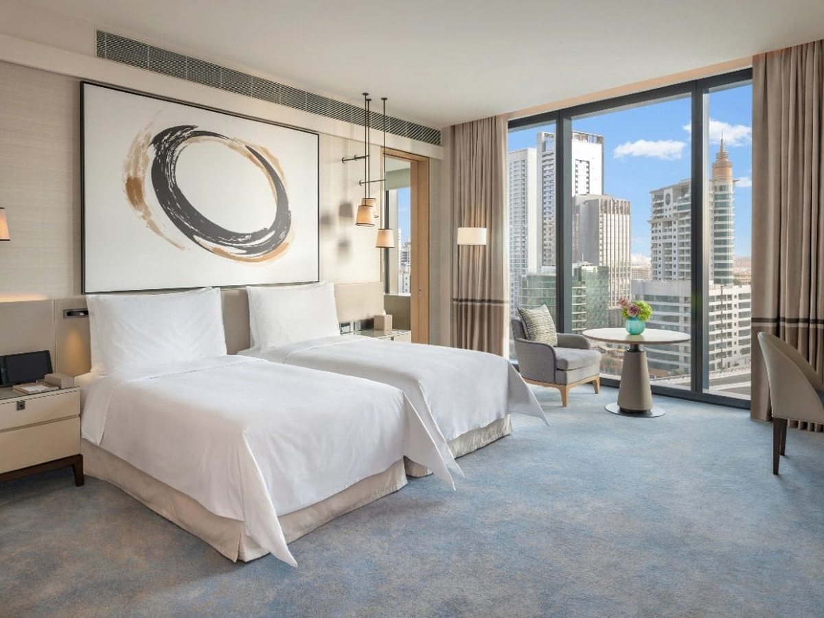 A room with two single beds in pale blue and cream, a small seating area and full-height windows over the Dubai Marina skyline.