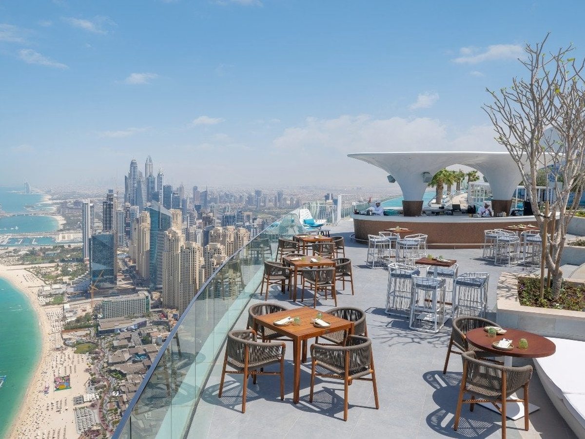A rooftop restaurant terrace with rope-and-timber chairs beside a glass balustrade, a sculptural bar canopy and the Dubai Marina skyline far below.
