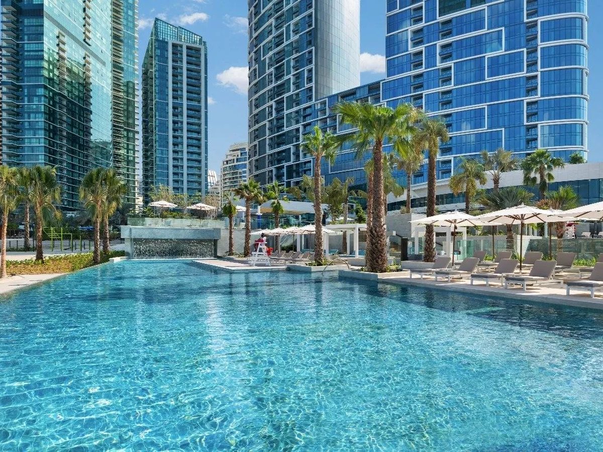 A large ground-level pool with a waterfall wall, ringed by palms, sun loungers and parasols, a red lifeguard chair and the resort towers behind.