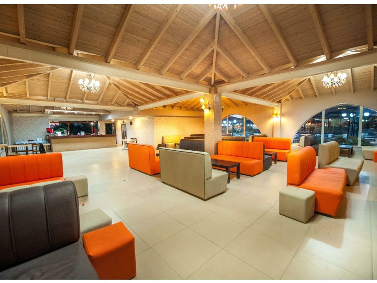 An open-plan indoor lounge with orange and grey modular sofas under a high timber vaulted ceiling and chandeliers, beside an arched bar.