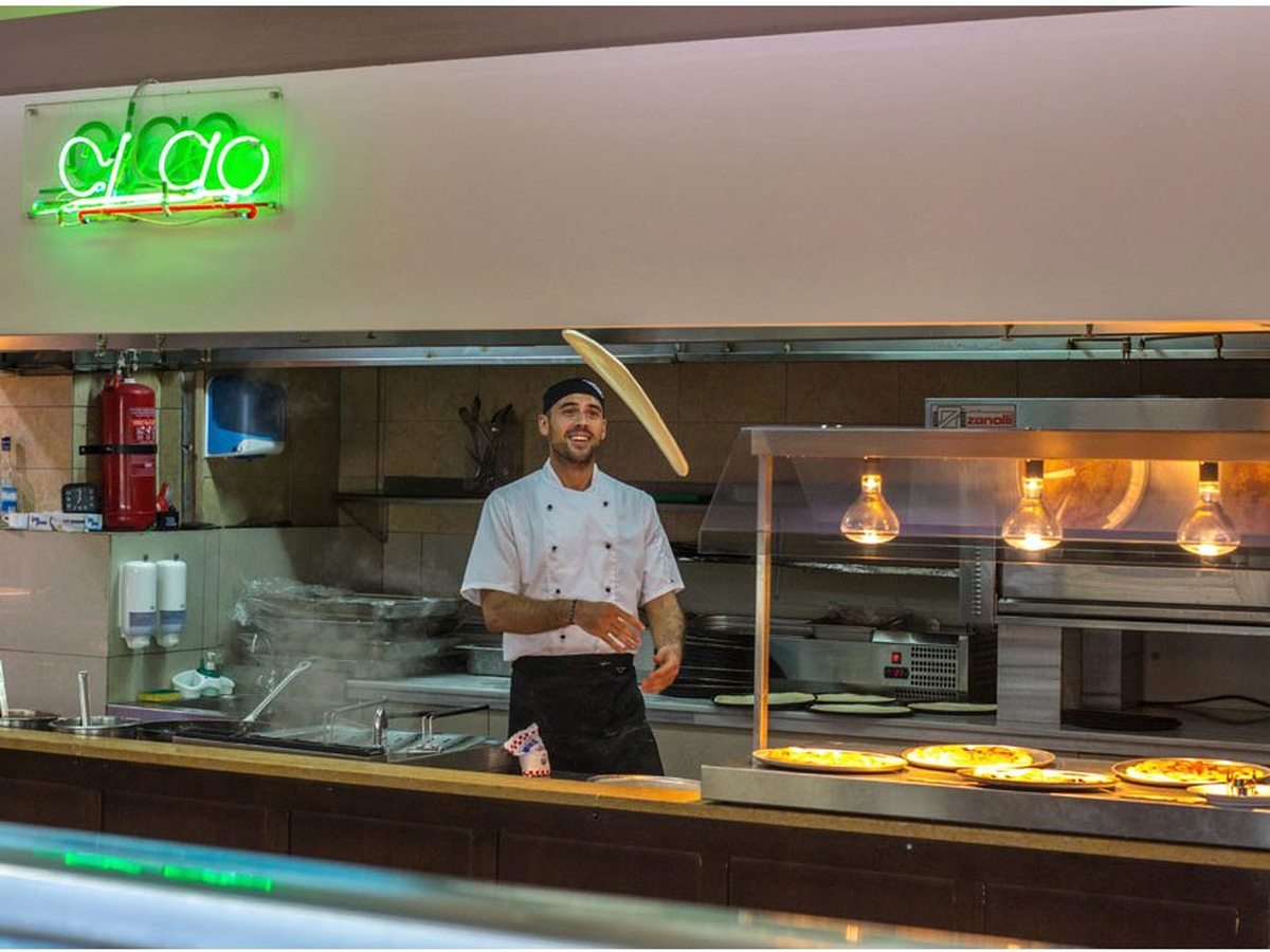 A chef working at a hot-food show-cooking counter under heat lamps, with a green neon sign above.