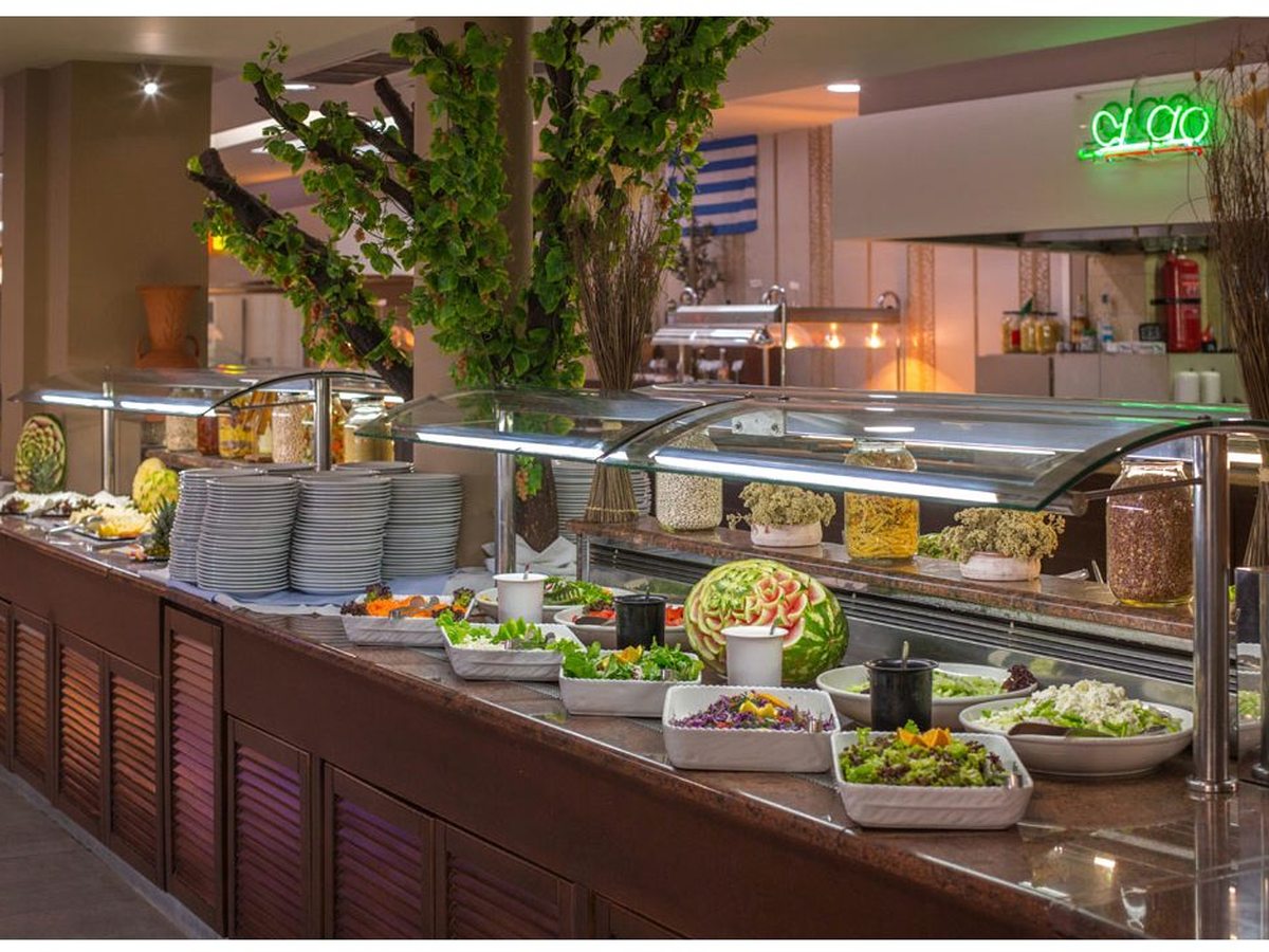 A long salad and cold buffet counter under glass guards with stacked plates, beside a decorative indoor tree.