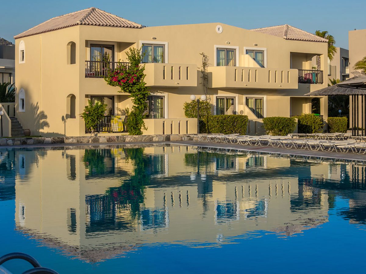 A freshwater pool reflecting a two-storey accommodation block with balconies, sun loungers and trimmed hedges alongside.