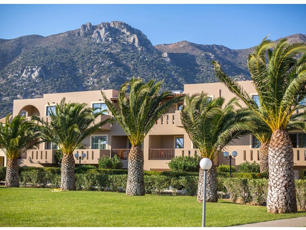 Two-storey garden accommodation buildings with balconies among palms and shrubs, with a rocky mountain behind under a clear sky.