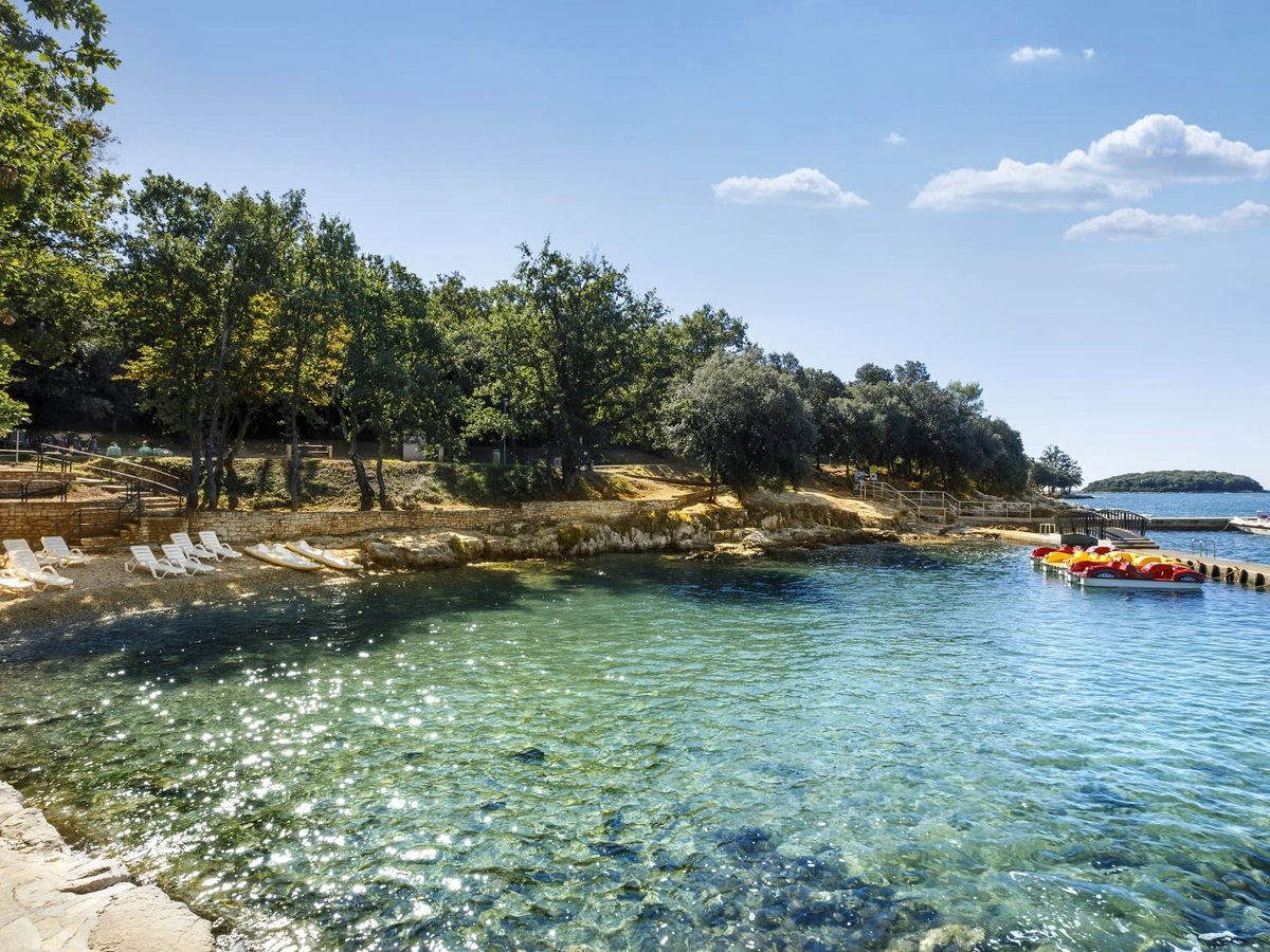 A rocky, tree-fringed cove with clear turquoise water and a stony shoreline, red and yellow pedal boats moored off.