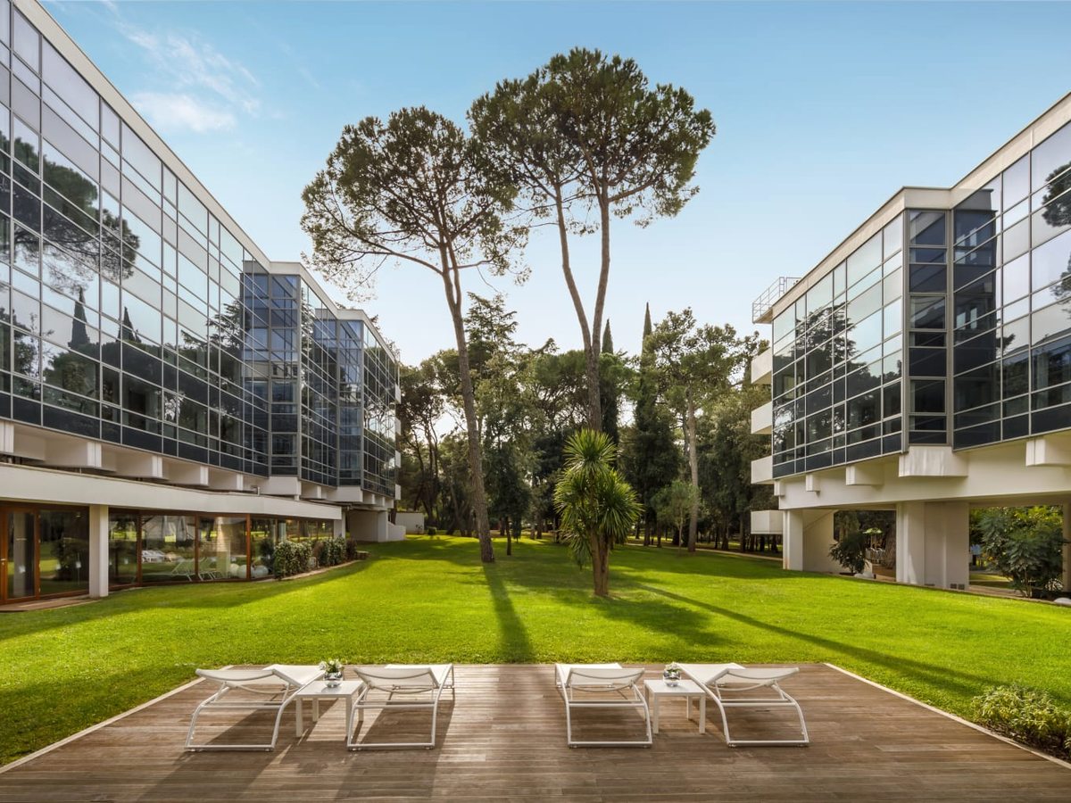A lawned courtyard between low glass-fronted room buildings, with sun loungers on a paved terrace and tall trees behind.