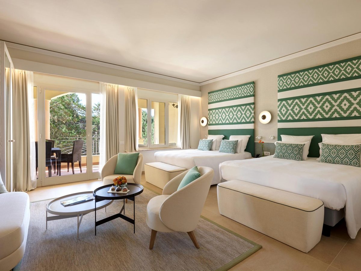 A family room with two large beds under geometric sage-green headboards, a small sitting area with armchairs, and a furnished terrace beyond.