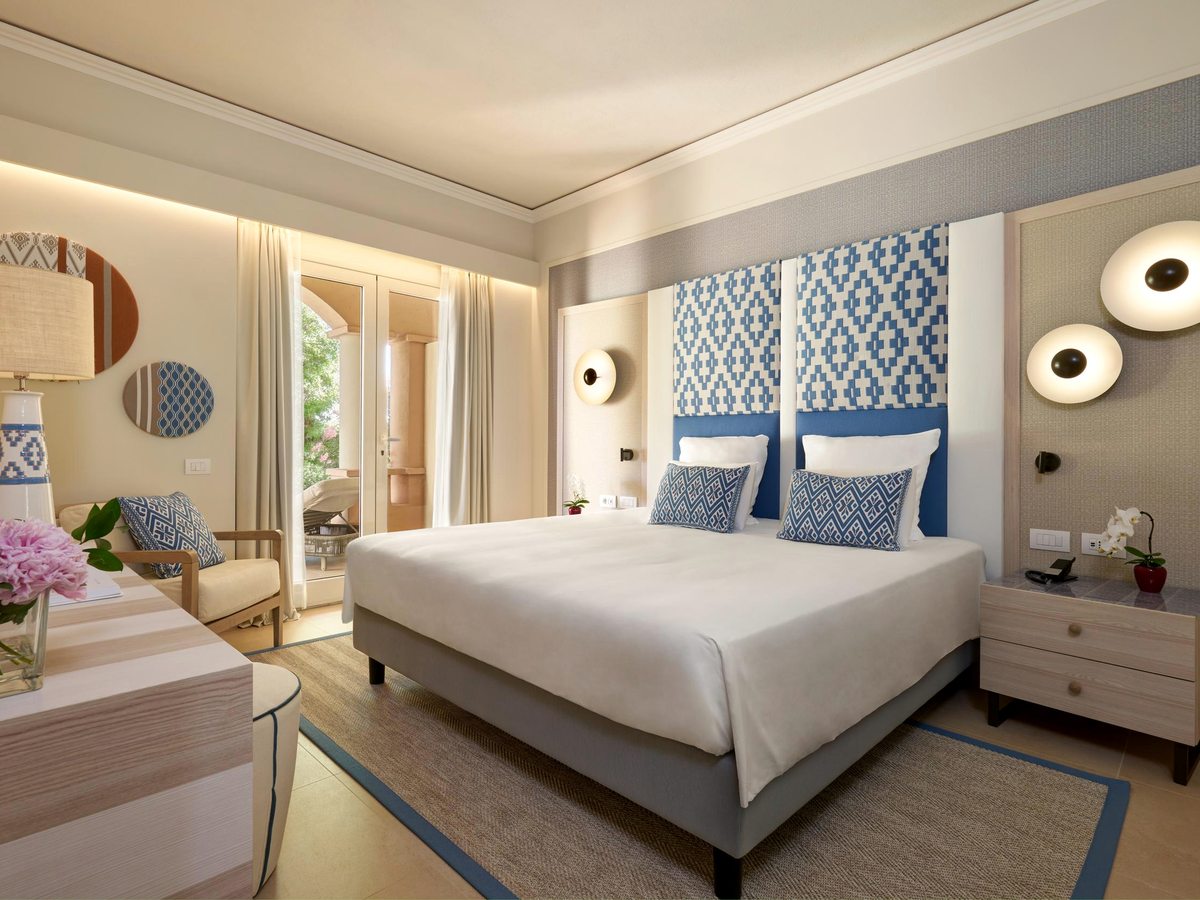 A double room with a blue patterned headboard, bedside tables, a vanity desk and armchair, and balcony doors letting in daylight.