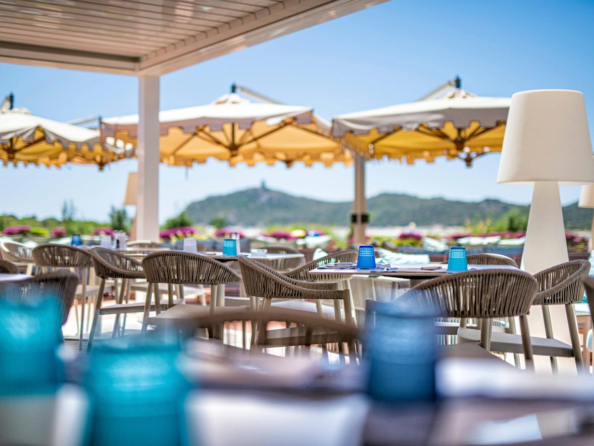 Outdoor restaurant tables under white parasols on a terrace, with grey chairs and a green hillside beyond.