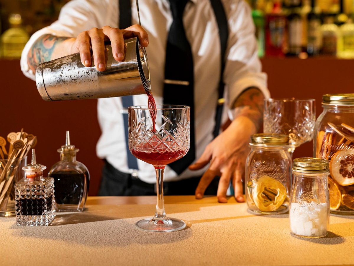 A bartender pouring a red cocktail into a stemmed glass at a bar counter lined with bottles and bar tools.