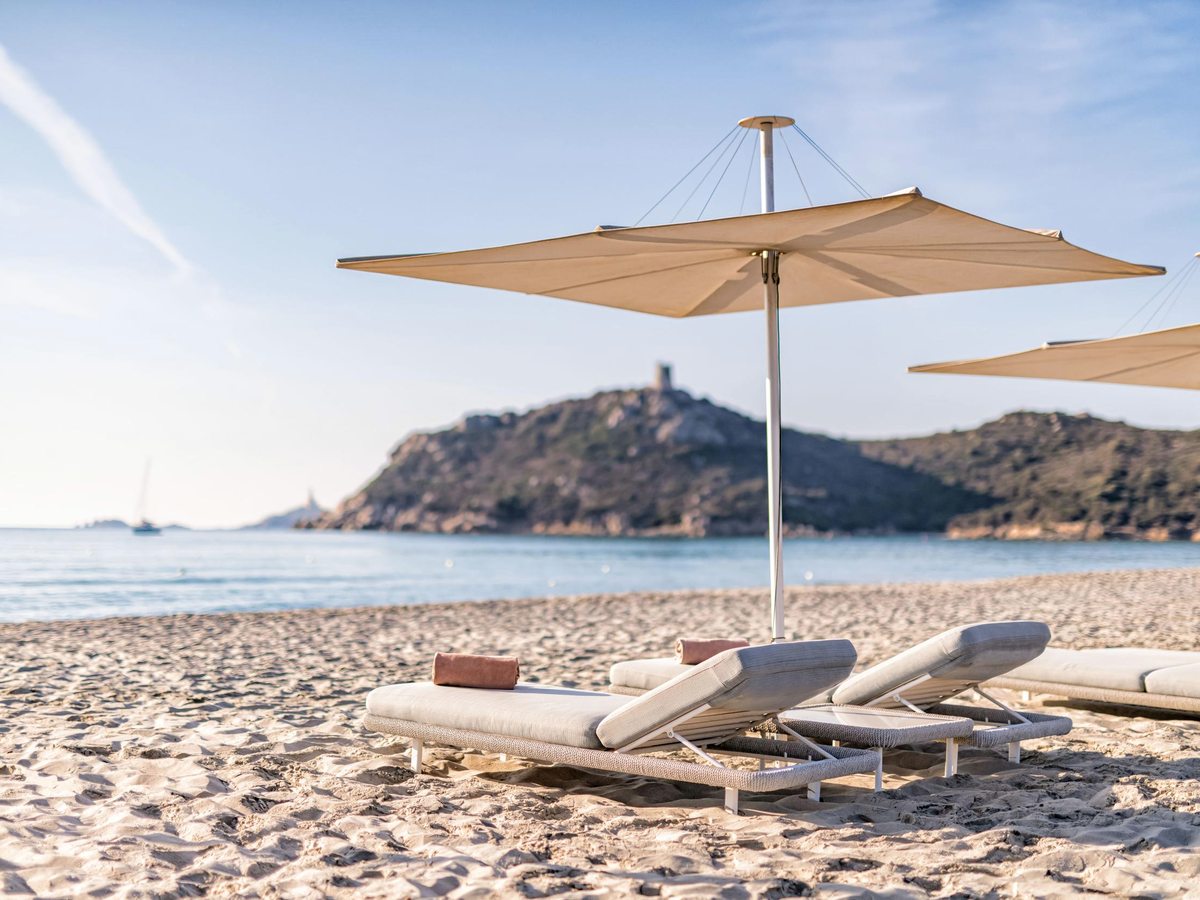 Sun loungers with cream parasols on the sand, the sea beyond and a stone watchtower on the hilltop headland.