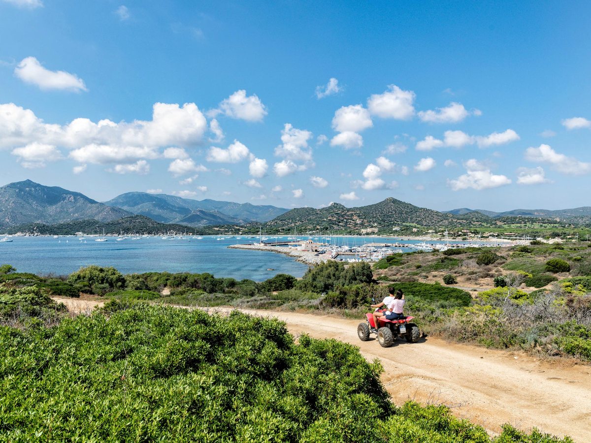 A quad bike on a dirt track through green coastal scrubland, with a wide bay and distant hills beyond.