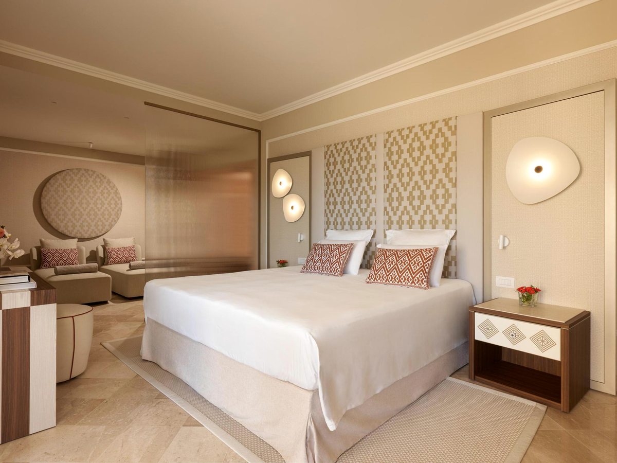 A suite with a king bed under a panelled headboard and an alcove sitting area with a sofa and lounge chairs.