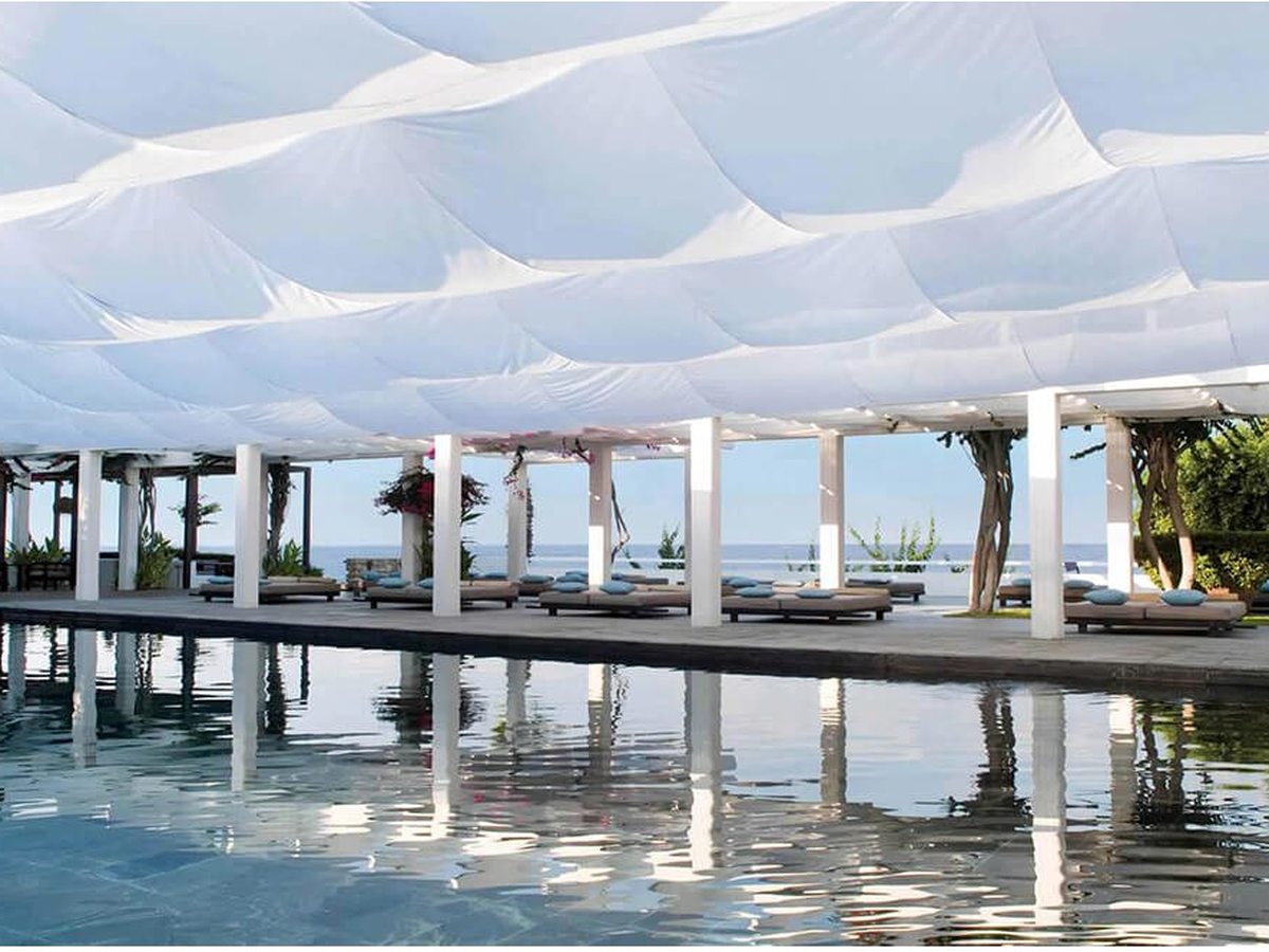 A freshwater pool beneath a rippling white fabric canopy on white columns, daybeds along the deck and the sea beyond.