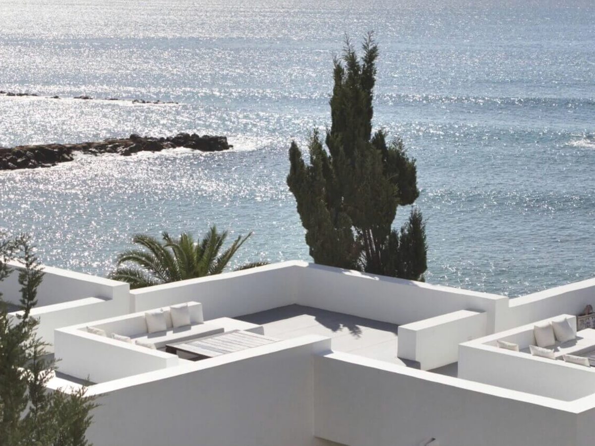 White cubic terraces with cushioned daybeds and a cypress tree, above a bright, sparkling sea.