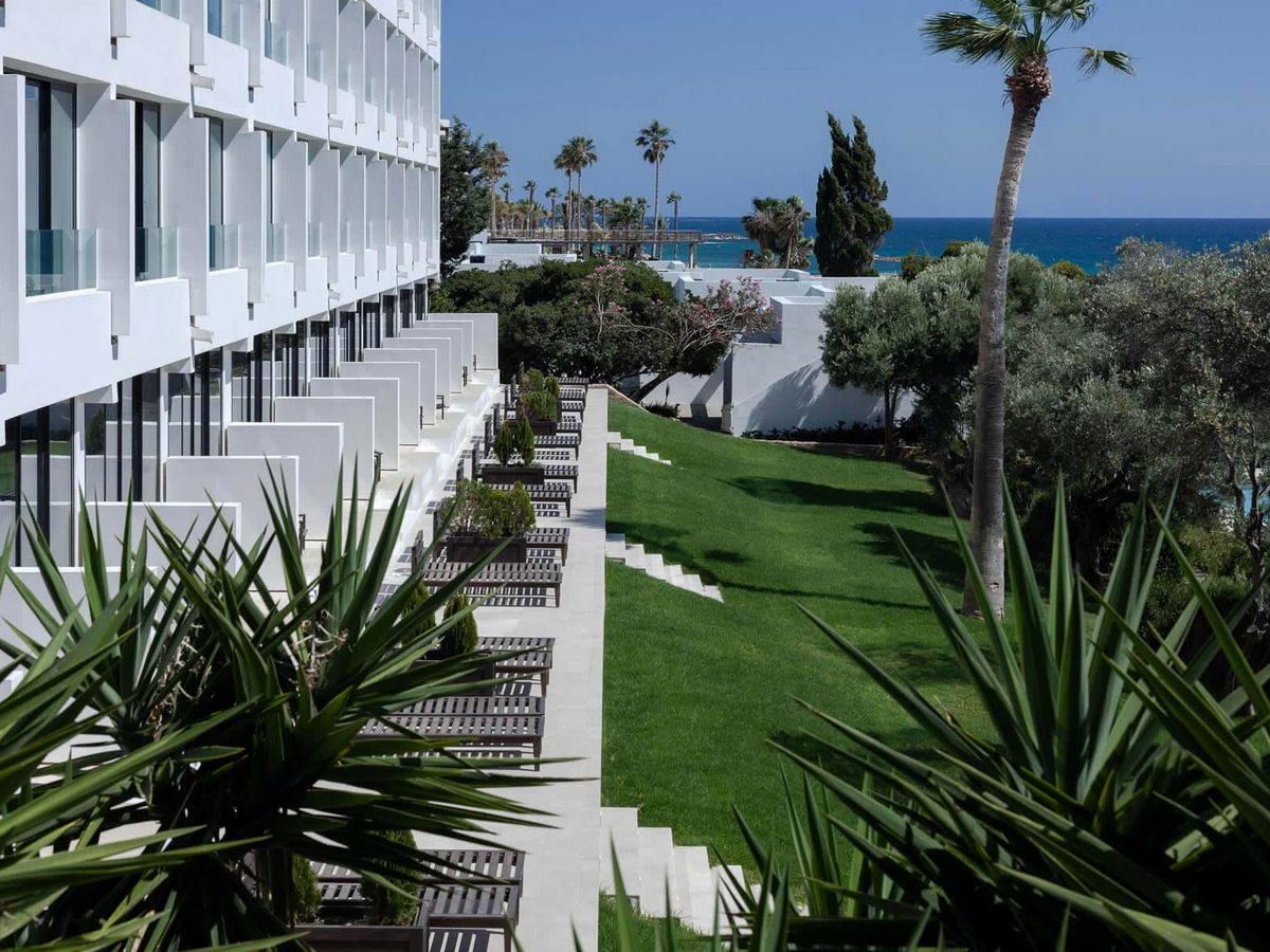 The white hotel's stepped room terraces lined with dark sunloungers, a marble stair and a lawn running down through palms and olive trees to the sea.