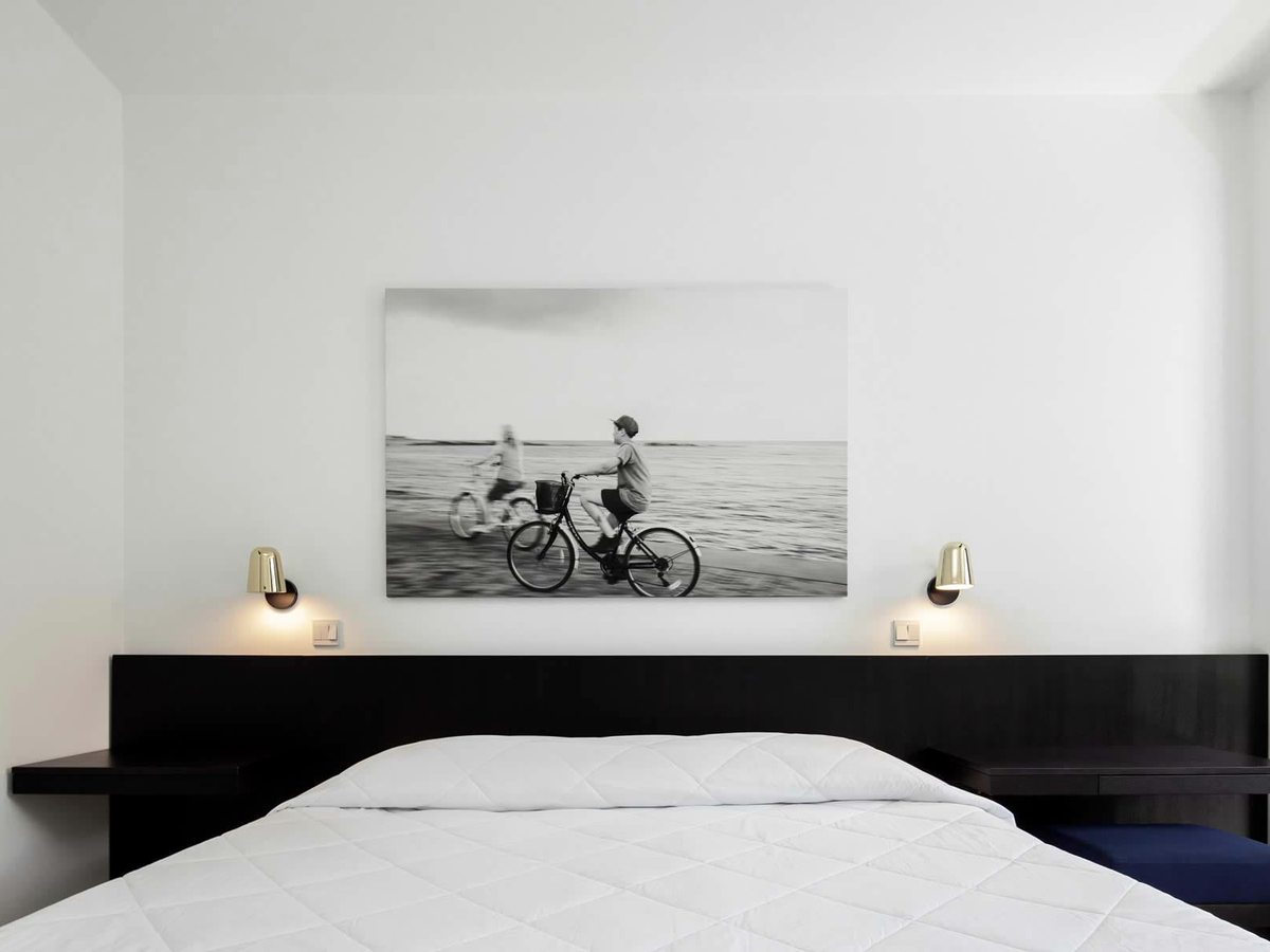 A double bed with a black timber headboard and brass reading lights, a large black-and-white photograph of cyclists on a beach hung above it.