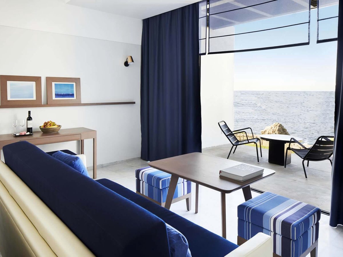 A suite living room with a navy sofa, blue striped stools and a console holding wine and fruit, opening through full-height glass to a sea-view terrace.