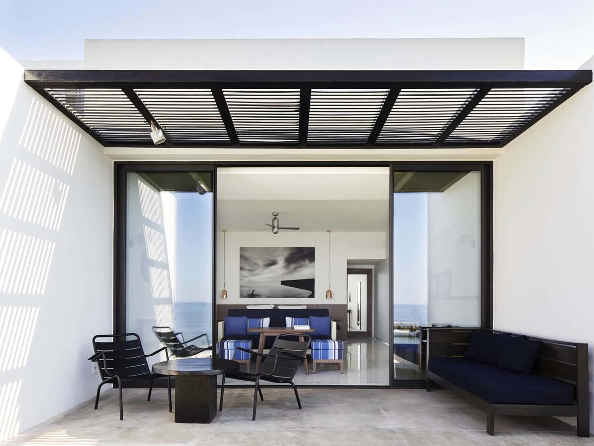 A suite terrace under a black slatted pergola with lounge chairs and a navy daybed, sliding glass doors back into the living room.