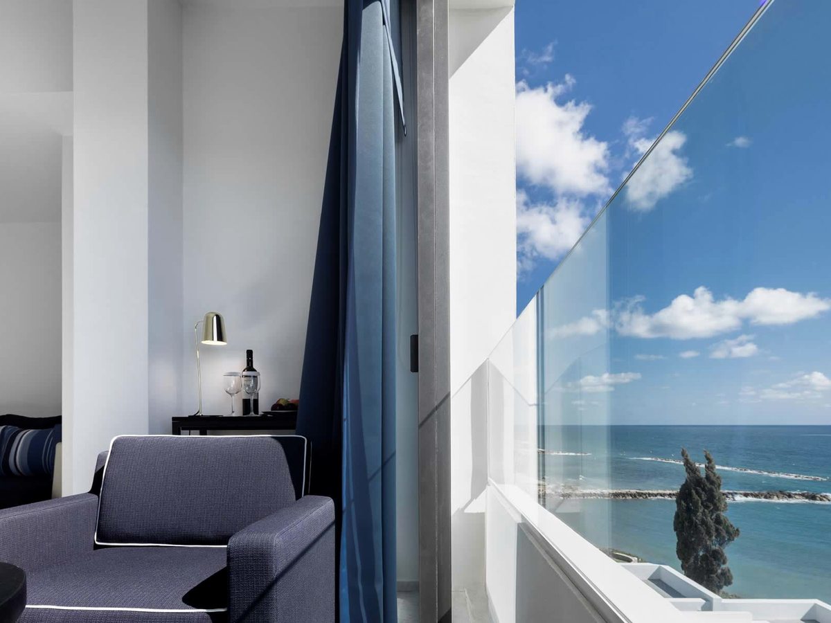 A room balcony with a grey armchair and a minibar set with wine glasses, a glass balustrade and the sea and breakwater beyond.