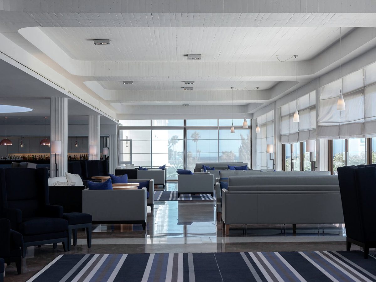 An open-plan lobby lounge with grey and navy armchairs, blue cushions and a striped rug, a bar counter along one wall and sea-view windows.