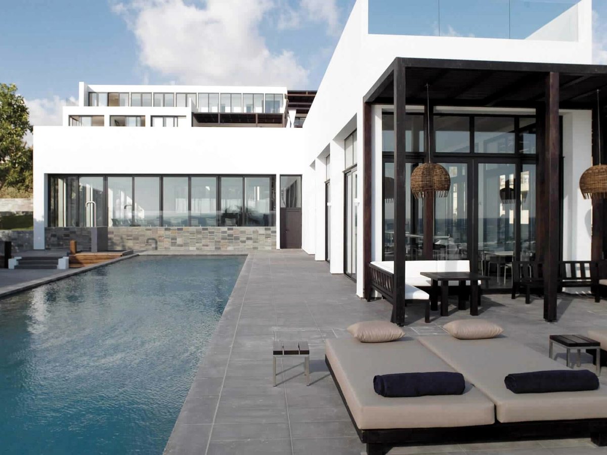 A pool beside the white hotel building with cushioned daybeds under a dark timber pergola.